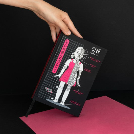 Mão segurando o caderno Squid Game com capa preta e ilustração temática.