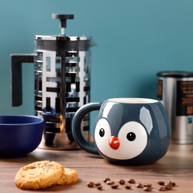 Caneca em formato de pinguim sobre uma mesa de cafe com bolachas e prensa francesa ao fundo.