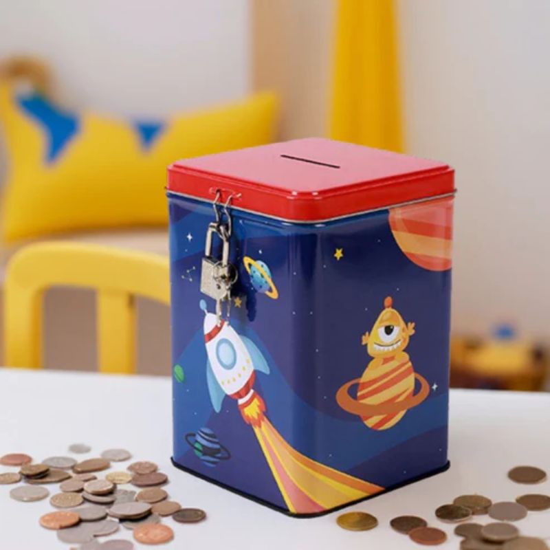um mealheiro retangular em metal com cadeado e desenhos infantis espaciais rodeado por moedas