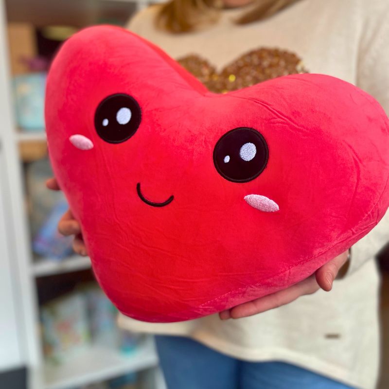 Almofada em forma de coração com rosto sorridente, segurada por uma pessoa. Perfeita para oferecer como presente romântico ou decoração aconchegante.