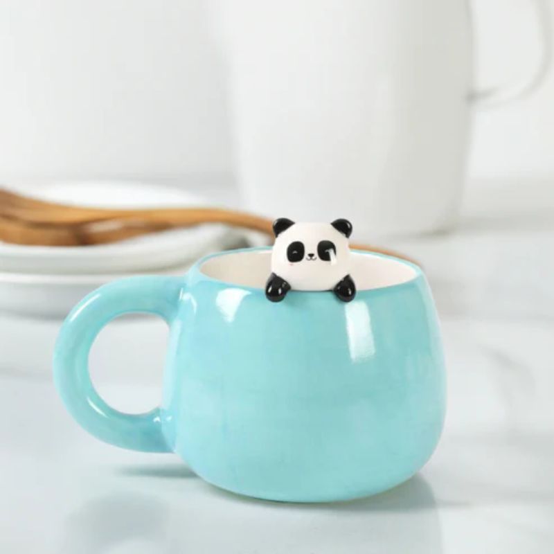 Caneca azul com figura de panda a espreitar do interior, sobre mesa com pratos e colheres ao fundo.
