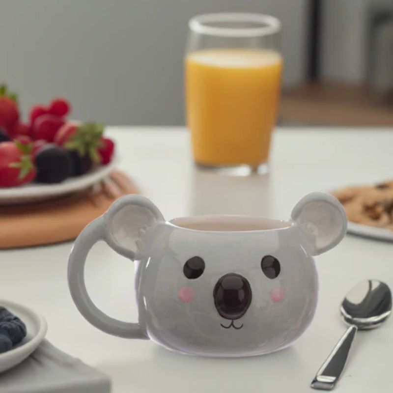 Caneca cinzenta em forma de coala com expressão adorável, sobre mesa de pequeno-almoço com fruta e sumo de laranja ao fundo.