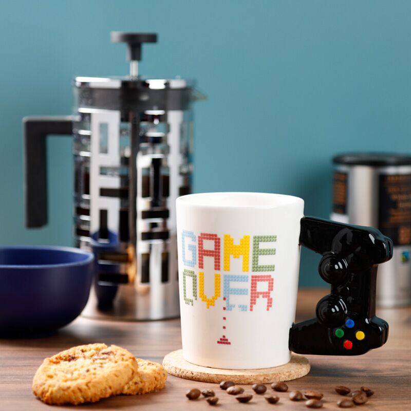 Caneca Game Over com pega em forma de comando numa mesa com café.