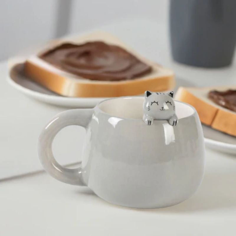 Caneca cinzenta com figura de gatinho a espreitar na borda, sobre mesa com torradas com creme de chocolate ao fundo.