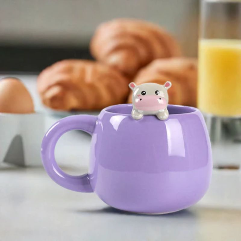 Caneca roxa com figura de hipopótamo a espreitar, sobre mesa de pequeno-almoço com croissants e sumo de laranja ao fundo.