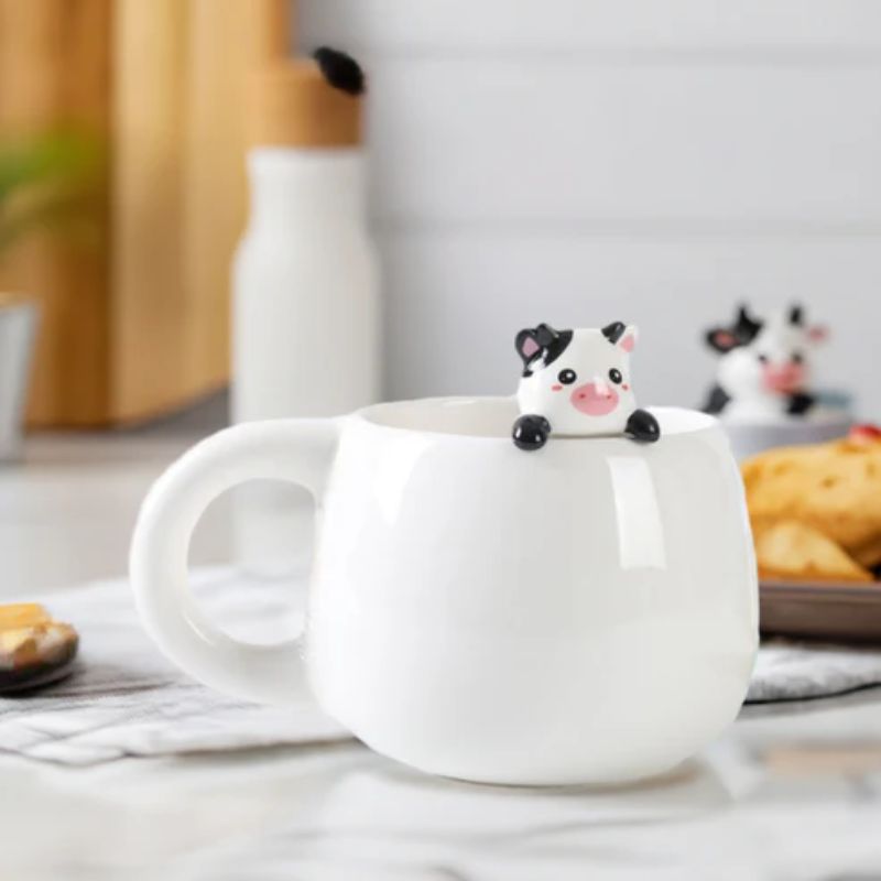 Caneca com vaquinha a decorar uma mesa de pequeno-almoço, com bolachas e frasco ao fundo.