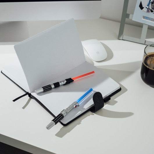 Duas canetas sabres de luz Star Wars posicionadas sobre um caderno aberto, com luzes LED apagadas.