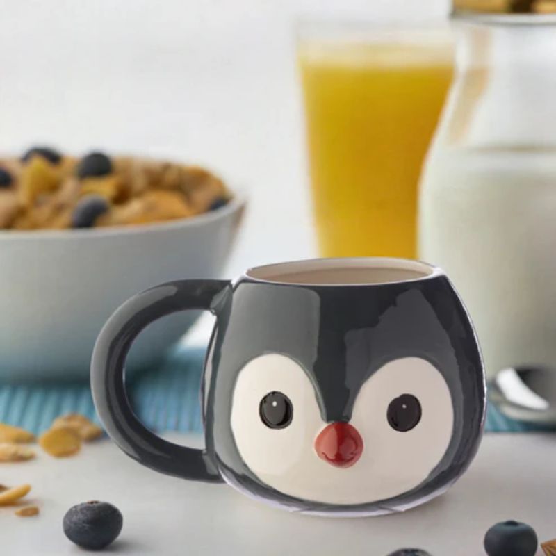 Caneca em forma de pinguim sobre mesa de pequeno-almoço, com sumo de laranja e cereais ao fundo.