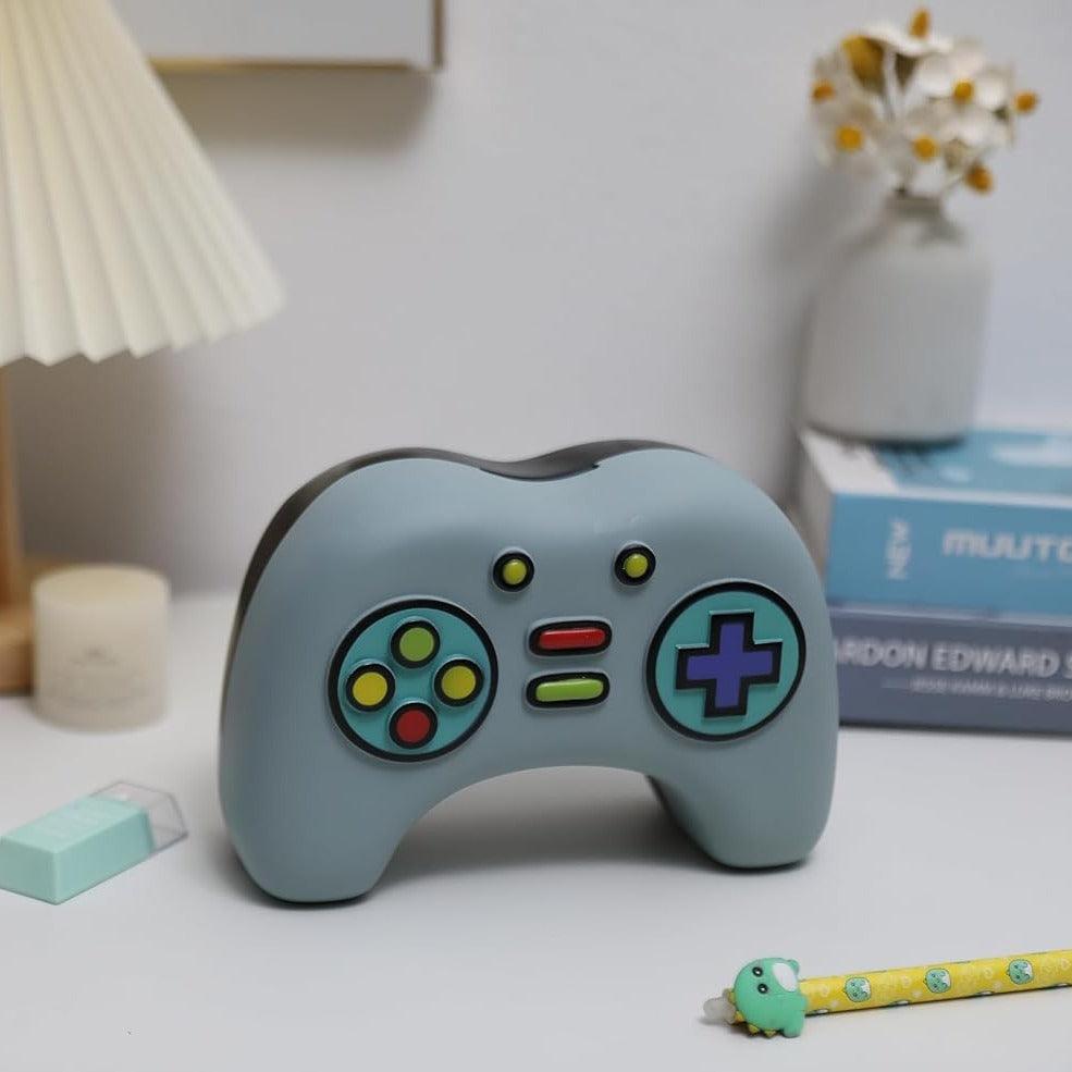 Mealheiro gamer em forma de comando colocado sobre secretária com livros e decoração.